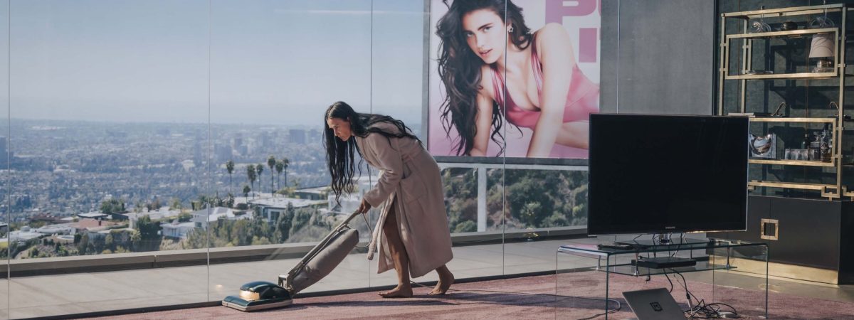 Demi Moore's character in THE SUBSTANCE vacuuming her apartment in the shadow of a billboard featuring her other self played by Margaret Qualley, from the film.